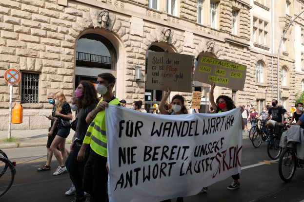 Eine Gruppe von Menschen marschiert auf einer Stadtstraße bei einer Protestaktion, einige halten Schilder und fahren Fahrräder, im Hintergrund ein historisches Gebäude mit Rundbogenfenstern, Säulen und Skulpturen.
