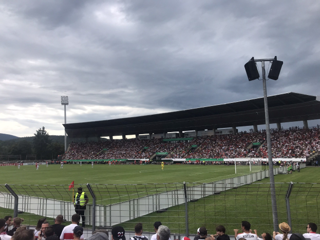 Große Zuschauermenge bei einem Fußballspiel in einem bewölkten, eingezäunten Stadion mit beleuchtetem Spielfeld, umgeben von Bäumen und Hügeln.