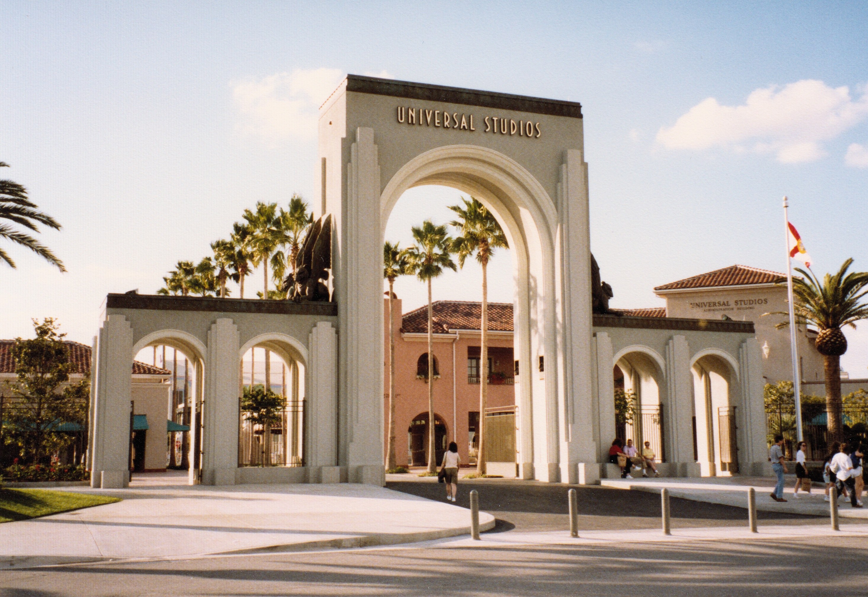 Eingang zum Universal Studios Hollywood mit einem Bogen mit Text, einer Statue, Menschen, Bäumen, Gebäuden, einem Fahnenmast und einem bewölkten Himmel.
