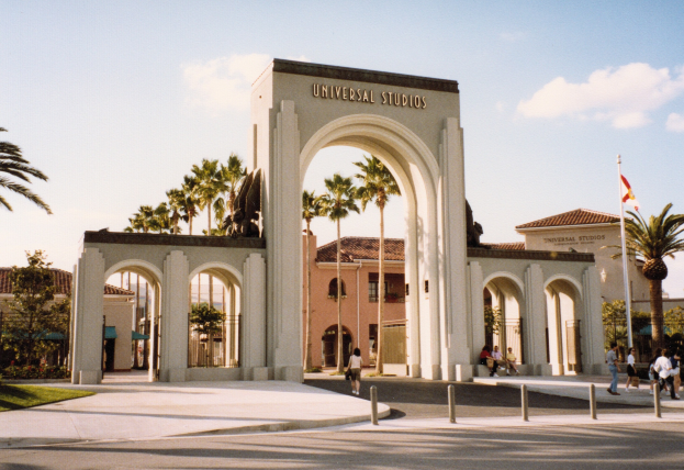 Eingang zum Universal Studios Hollywood mit einem Bogen mit Text, einer Statue, Menschen, Bäumen, Gebäuden, einem Fahnenmast und einem bewölkten Himmel.