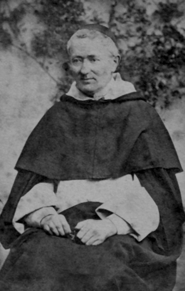 Altes Schwarz-Weiß-Foto eines Mannes in Priesterrobe, der vor Bäumen sitzt, mit schwarzer Robe, weißem Kragen und Manschetten, die Hände zum Gebet gefaltet, von warmem Licht beleuchtet mit friedlichem Gesichtsausdruck.