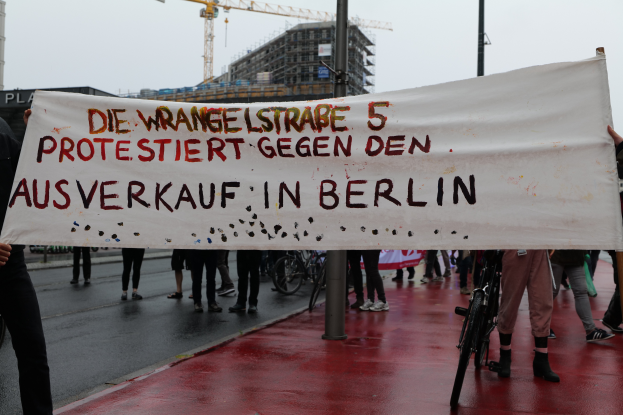 Eine Gruppe von Menschen steht auf der Straße und hält eine Plakette mit der Aufschrift "Die Wrangellstraße 5 protestiert gegen den Ausverkauf in Berlin". Im Hintergrund sind Fahrräder, Stäbe, Gebäude und ein Kran zu sehen, öber ihnen der Himmel.