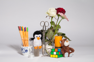 Ein weißer Tisch mit einer Vase voller bunter Blumen, einem Teddybär, einem Becher voller Stifte, einem Spielzeugpinguin, einer Kerze, einer Schere und einem Rubik's Cube.