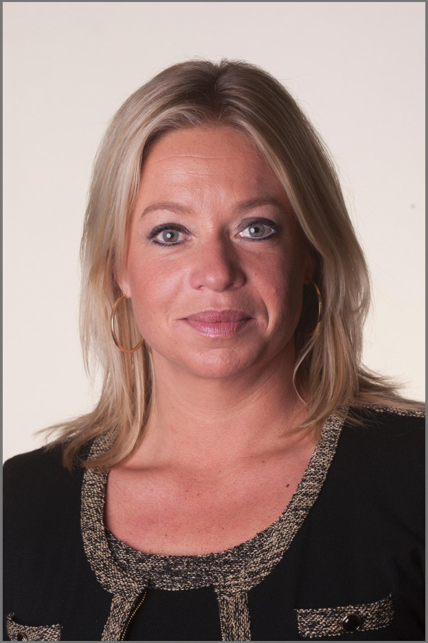 Eine Frau mit blondem Haar in einem ordentlichen Dutt, die ein schwarzes Kleid und goldene Ohrringe trägt und einen warmen und selbstbewussten Ausdruck hat.