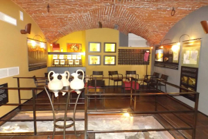 Ein Museumsraum voller Tische, Stühle, Geländer, Vasen, Texttafeln, Wandrahmen, Lampen und diverser Ausstellungsstücke.