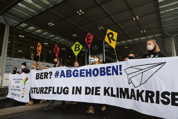 Eine Gruppe maskierter Personen mit einem Banner "Sturzflug in die Klimakrise" vor einem Glasgebäude mit Säulen, beleuchtet von Deckenlampen.