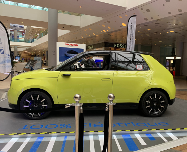 Gelber wasserstoffbetriebenes Elektroauto vor einem Gebäude mit Absperrpollern, Bannern, Menschen, Schilden, einer Topfpflanze, Deckenleuchten und Ventilatoren.