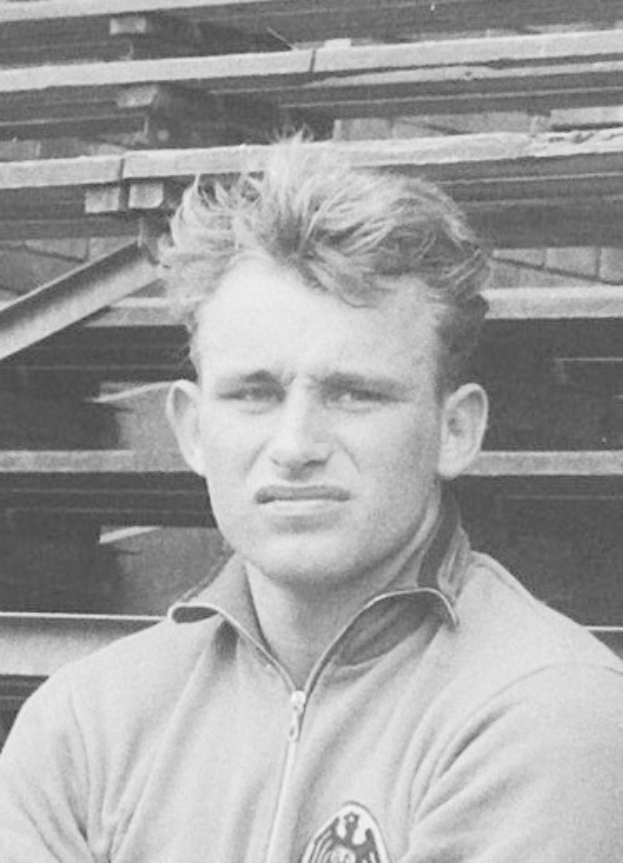 Schwarzes und weißes Foto eines Mannes in einer Jacke, der vor gestapelten Holzplanken steht und für den legendären deutschen Fußballspieler gehalten wird.