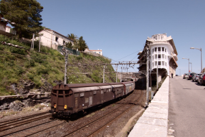 Güterzug fährt auf Gleisen neben einem Gebäude mit parkenden Autos auf einem Bahnsteig auf der rechten Seite, vor einer Kulisse aus städtischen Gebäuden, Bäumen, Strommasten, Drähten und einem klaren blauen Himmel.