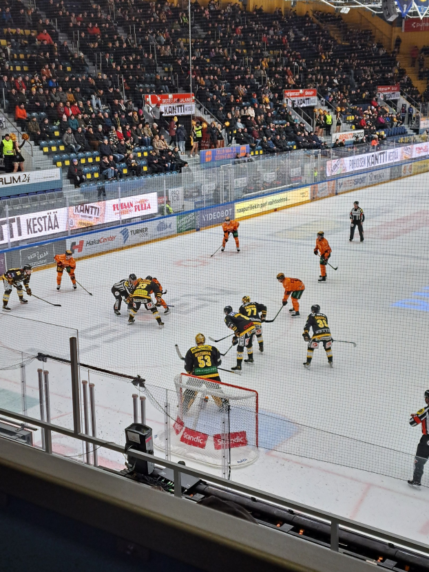 Eine Gruppe von Menschen, die Hockey auf einem Eisring mit einem Netz und einer umgebenden Absperrung spielen, Zuschauer sitzen im Hintergrund und Banner mit Text, unter hellen Stadionlichtern.