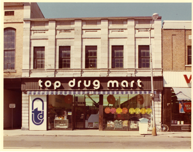 Eckladen namens "Top Drug Mart" mit Fenstern, einem Schild, einem Straßenschild, einem geparkten Fahrrad, einem Müllcontainer und einem bewölkten Himmel.