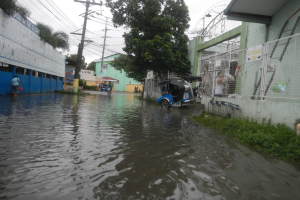 Eine überflutete Straße mit einem geparkten Motorrad in der Mitte, Gebäude, Bäume und Strommasten auf beiden Seiten und einige Menschen und Fahrzeuge im Hintergrund.