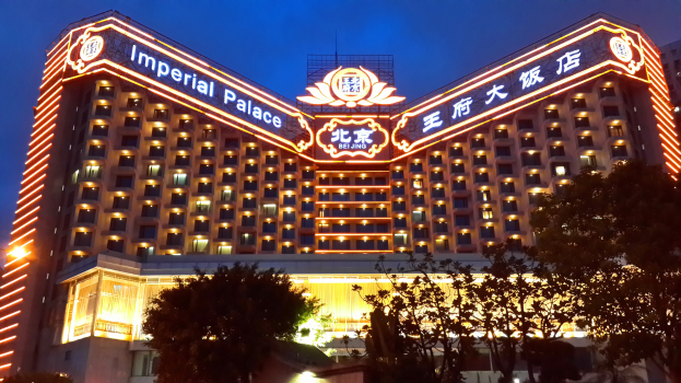 Großes Gebäude des Kaiserpalast-Hotels in Peking mit vielen Fenstern und Lichtern, einem Namenstafel, Bäumen im Vordergrund und einem klaren blauen Himmel.