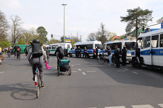 Eine Gruppe von Menschen auf Fahrrädern auf einer Straße mit Polizeiwagen, anderen Fahrzeugen, Bäumen und Gebäuden im Hintergrund.