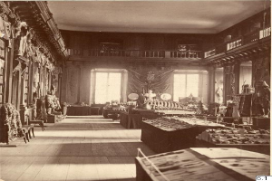 Ein altes Schwarz-Weiß-Foto eines Raumes voller Tische, Stühle, Skulpturen an den Wänden, Fenstern, Vorhängen, Geländern und anderen Gegenständen mit der Aufschrift "The Museum of Fine Arts, London" unten.