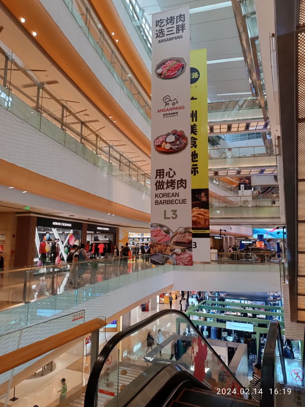 Einblicke in ein Einkaufszentrum mit einer Rolltreppe in der Mitte, Menschen, die herumgehen, einige mit Handtaschen, und Schilder, die von der Decke hängen, mit Geschäften im Hintergrund.