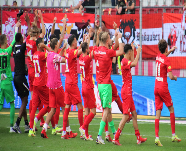 Eine Gruppe von Fußballspielern feiert ein Tor auf einem Feld, einige halten Flaschen und andere klatschen, mit einem Zaun, Bannern, Metallstangen, Stühlen und einem Dach mit Lampen im Hintergrund.