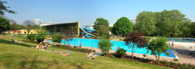 Großer Swimmingpool umgeben von grünem Gras, Bäumen, Menschen im und um das Wasser, mit Gebäuden und einem klaren blauen Himmel im Hintergrund.