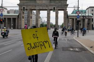 Eine Gruppe von Menschen auf Fahrrädern fährt vor dem Brandenburger Tor in Berlin, Deutschland, vorbei, wobei sie Helme tragen und einer ein gelbes Schild hält, mit Laternenpfählen, Verkehrszeichen, Gebäuden, Bäumen und einem klaren blauen Himmel im Hintergrund.