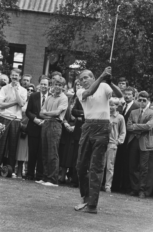 Ein Mann schwingt einen Golfschläger vor einer Menge, mit einem Gebäude und Bäumen im Hintergrund, dargestellt in Schwarz-Weiß.