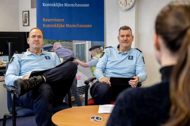 Zwei Polizeibeamte sitzen an einem Tisch mit einer Frau, umgeben von Dokumenten und Gegenständen, mit einem Banner, einer Uhr und Foto Rahmen an der Wand dahinter und einem weiteren Tisch mit einem Monitor und Zubehör links daneben.