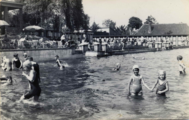 Schwarz-weißes Foto von Kindern, die in einem Swimmingpool mit Menschen, Sonnenschirmen, Gebäuden und Bäumen im Hintergrund und einem klaren Himmel spielen.