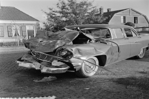 Ein Schwarz-Weiß-Foto eines schwer beschädigten Autos am Straßenrand mit eingedellter Motorhaube und zersplitterten Fenstern vor dem Hintergrund von Gebäuden, Bäumen und einem klaren Himmel.
