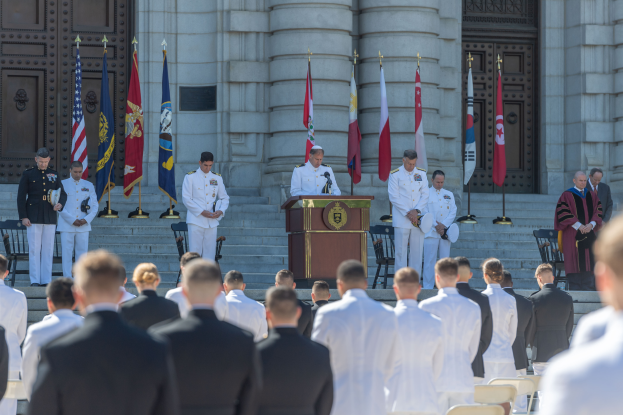Eine Gruppe von Männern in weißen Uniformen steht vor einem Gebäude mit Säulen und Türen, umgeben von einem Podium mit Mikrofon und Mikrofonständer, mit Flaggen und Stühlen im Hintergrund, was an eine Abschlussfeier erinnert.