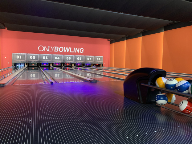 Eine Bowlingbahn mit Bowlingkugeln auf der rechten Seite, Pins in der Mitte und einer Wand mit Text im Hintergrund, unter einer sichtbaren Decke.