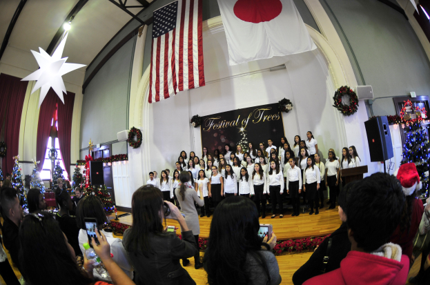 Eine Gruppe von Menschen auf einer Bühne vor einem Publikum mit geschmückten Weihnachtsbäumen, einem Podium, Lautsprechern, einer Texttafel, Vorhängen, Flaggen und Deckenleuchten.