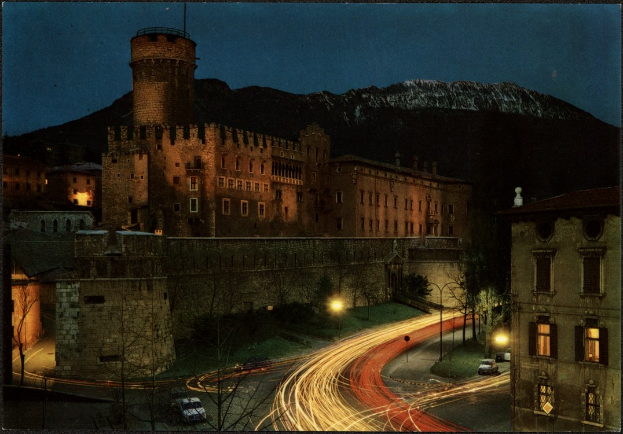 Ein bei Nacht beleuchtetes Schloss, das in einer städtischen Umgebung mit umliegenden Gebäuden, Straßeninfrastruktur, Fahrzeugen, Bäumen, fernen Hügeln und einem sternenklaren Himmel liegt.