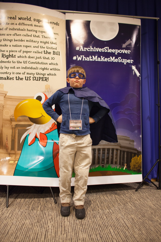 Ein junger Junge in einem Superheldenkostüm steht vor einem Banner mit Text und einem Vogelbild, mit einem Vorhang im Hintergrund; er trägt Schuhe.