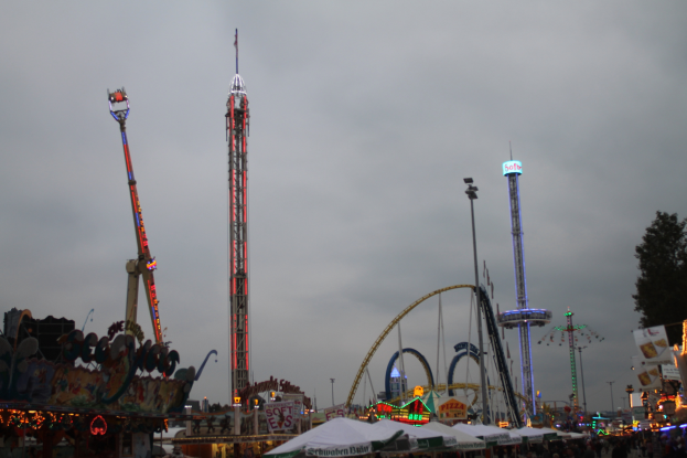 Eine Menschenmenge versammelt sich um einen hell erleuchteten Fasching bei Nacht, mit beleuchteten Fahrgeschäften, Polen und Bäumen unter einem bewölkten Himmel.