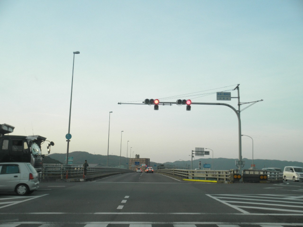 Ein Auto auf einer Autobahn an einer Kreuzung mit einer Ampel, umgeben von Fahrzeugen, einem Fußgänger, Geländern, Laternen, Verkehrszeichen, Straßenschildern, Gebäuden, Hügeln und einem klaren blauen Himmel.