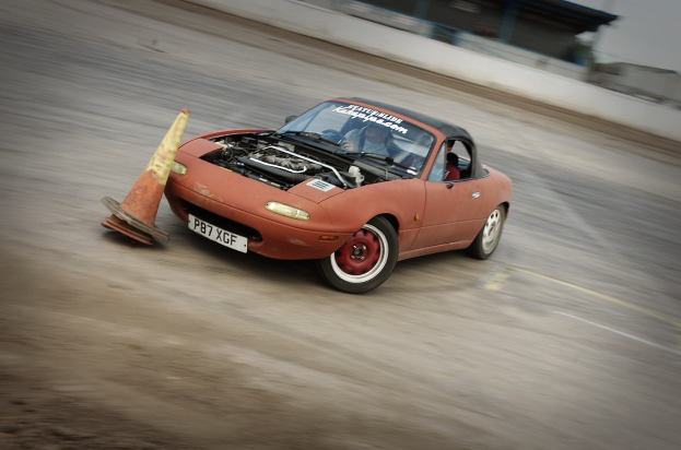 Eine Person, die ein rotes Auto auf einer Rennstrecke fährt, mit einem Verkehrskegel vor ihnen und einem unscharfen Hintergrund, der hohe Geschwindigkeit andeutet.