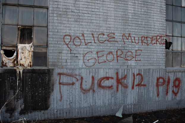 Graffiti auf einer Gebäudewand mit der Aufschrift "Polizeimord George Fuck 12 P19" und Fenstern und verstreuten Gegenständen davor.