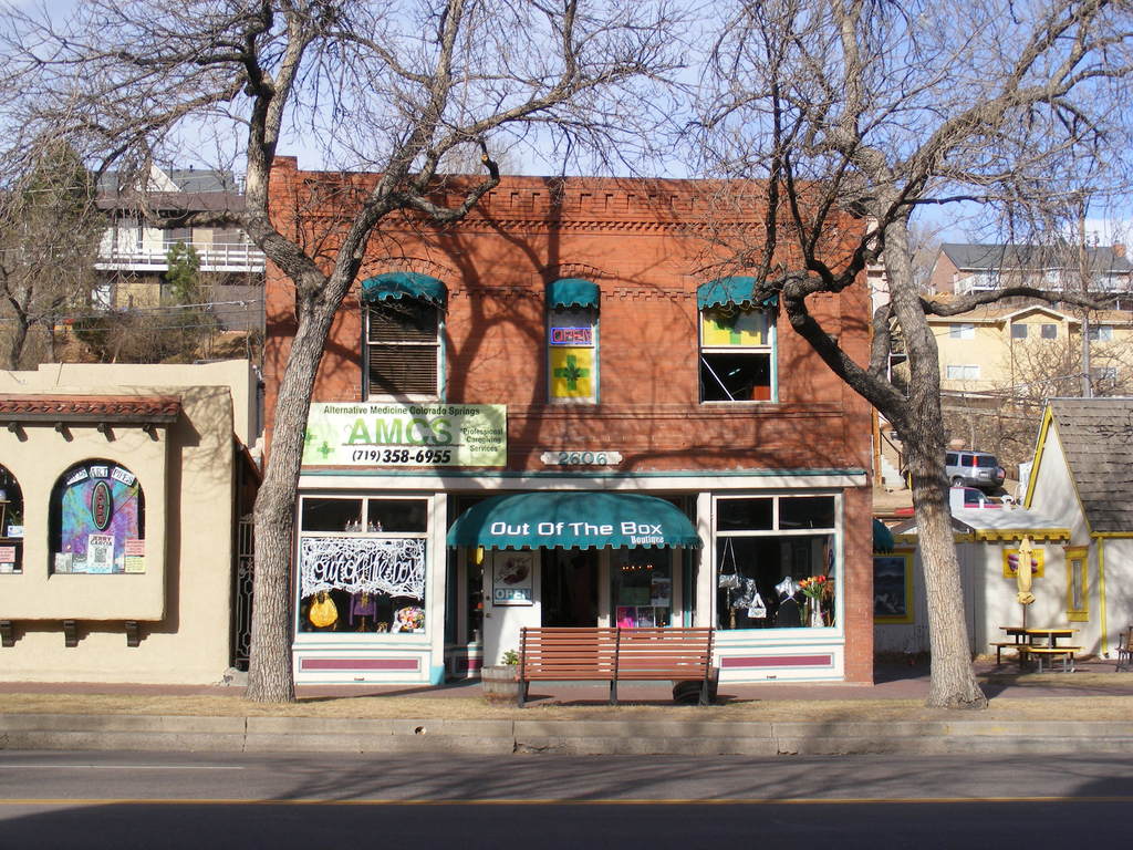 Eine Stra√e im Vordergrund mit Geb√§uden, B√§nken, B√§umen, einem Fahrzeug und dem Himmel im Hintergrund.