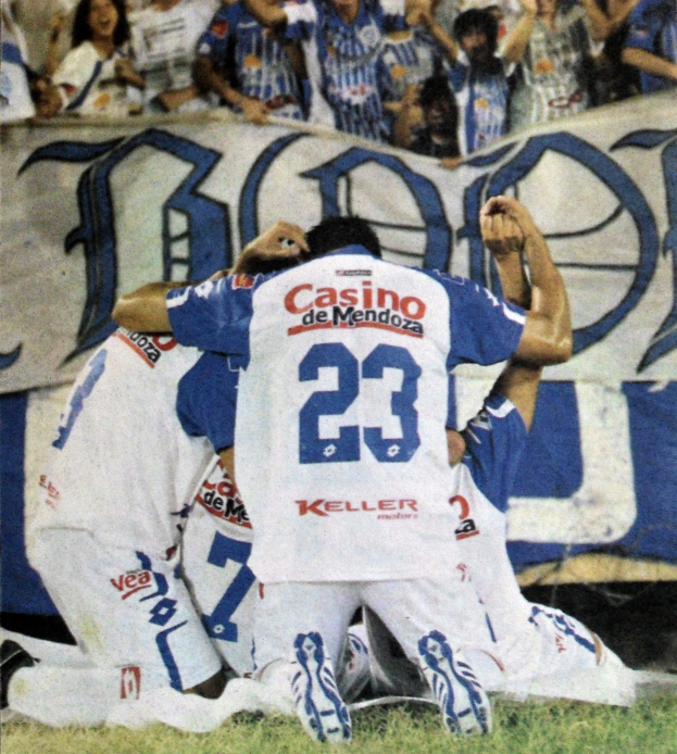Eine Gruppe von Fußballspielern feiert ein Tor auf dem Feld mit einer jubelnden Menge und einem Banner im Hintergrund.