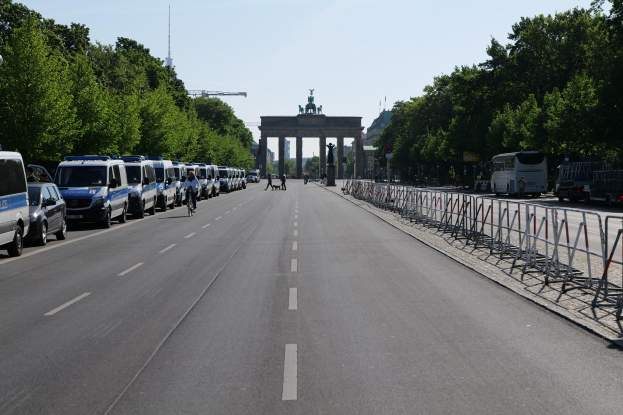 Lange Reihe von Polizeiautos auf der Straße vor dem Brandenburger Tor in Berlin, Deutschland, mit Menschen auf Fahrrädern und auf der Straße, Barrieren, Bäumen, einem Bogen mit Statuen im Hintergrund und sichtbarem Himmel.