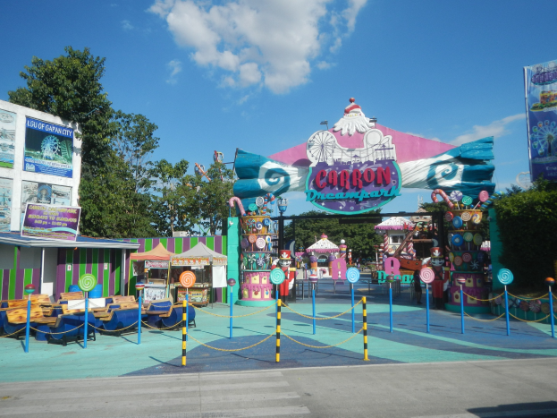 Karussell am Eingang eines Parks mit Straße, eingezäuntem Bereich, Buden, Banner, Bäumen und bewölktem Himmel im Hintergrund.