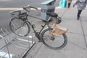 Ein grünes und braunes Fahrrad, das an der Seite einer Straße neben einem Müllcontainer geparkt ist, mit einigen Gegenständen daran befestigt, während Fahrzeuge auf der Straße vorbeifahren und Menschen auf dem Gehweg im Hintergrund zu sehen sind.
