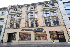 Ein Schaufenster namens "Schuhe & die Stadt" mit Schaufensterpuppen in Kleidern, Topfpflanzen, einem Schild, einer Stra├čenlaterne und Dr├Ąhten unter einem bew├Âlktem Himmel.