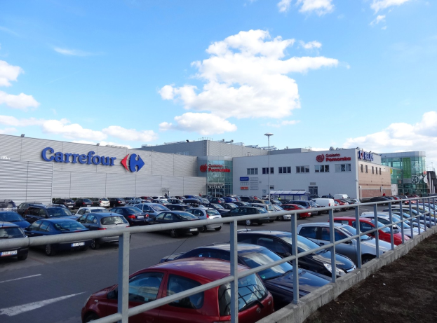Großer Autohandel mit parkenden Fahrzeugen vor einem städtischen Hintergrund mit Straßenlaternen, Schildern und vorbeifahrenden Kraftfahrzeugen unter einem bewölkten Himmel.