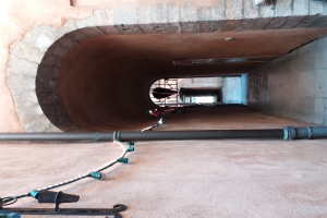 Ein großes Rohr in der Mitte eines Tunnels mit einer Wand auf der rechten Seite, einer Leiter im Hintergrund und ein paar Gegenständen auf der linken Seite.