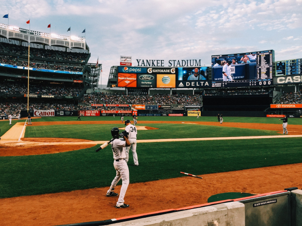 Baseballspiel im Yankee Stadium mit Spielern auf dem Feld und Zuschauern in den Rängen, umgeben von Stadionmerkmalen wie Zäunen, Fahnen, Anzeigetafeln, einem Display und Deckenleuchten unter einem bewölktem Himmel.
