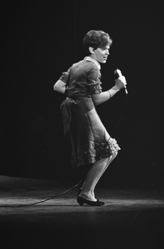 Eine Frau in einem Kleid steht auf einer Bühne, hält ein Mikrofon, trägt Schuhe, in einem Schwarz-Weiß-Bild.