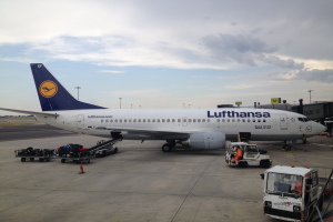 Ein Lufthansa-Airbus A320-200 steht am Frankfurter Flughafen, umgeben von verschiedenen Bodenfahrzeugen, Wagen, Absperrgittern, Polen und Texttafeln unter einem bewölkten Himmel.
