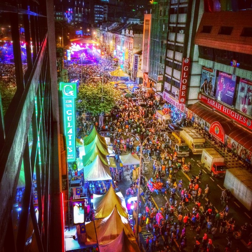Eine große Menge versammelt sich auf einer Straße, umgeben von Zelten, Geschäften, Gebäuden und Bannern, mit zahlreichen Lichtern im Hintergrund.