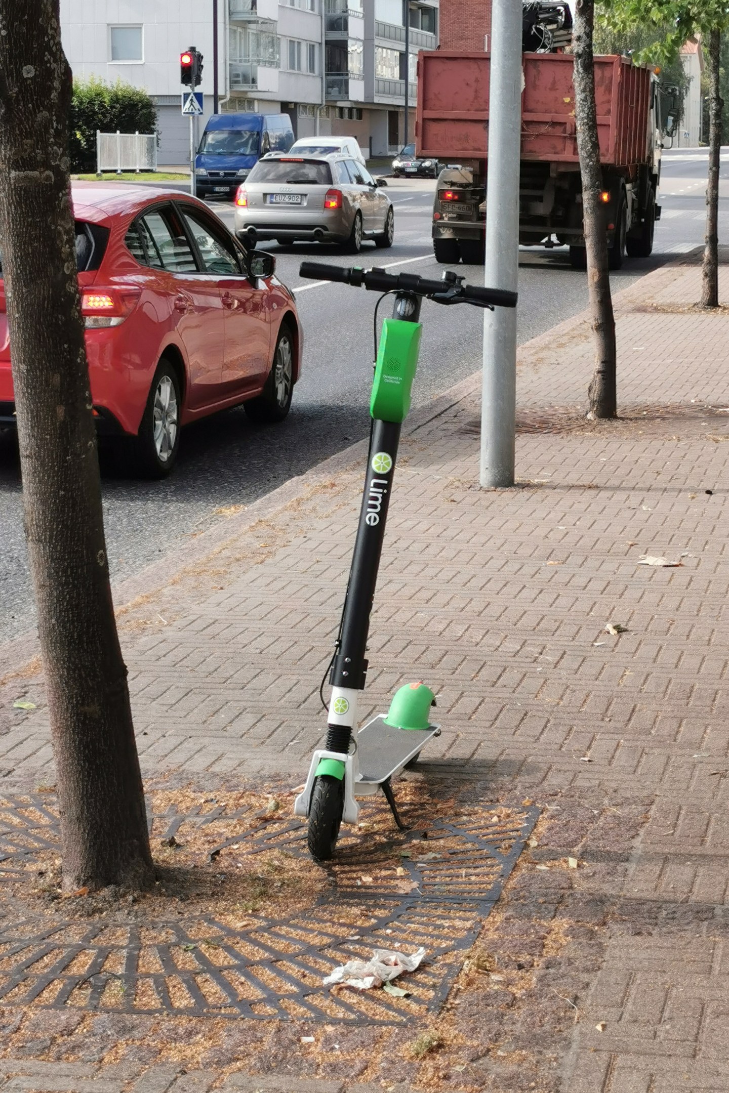 Elektrischer Roller auf einer Straße neben einem Baum geparkt, mit Fahrzeugen und Gebäuden im Hintergrund.
