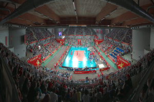 Ein großes Hallenstadion voller Zuschauer bei einem Volleyballspiel mit Bannern, Bildschirmen, Deckenleuchten und Geländern unten.
